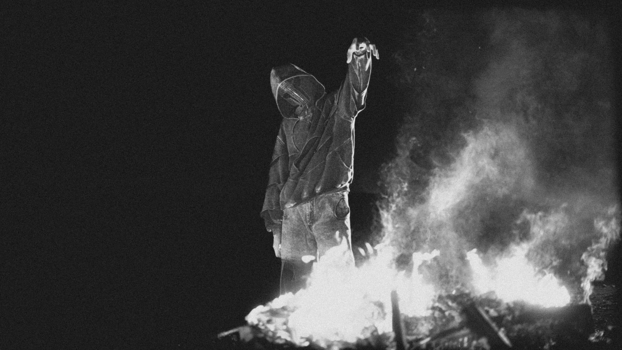 DRACU, hooded, one arm raised, standing over a fire at night